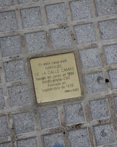A close-up of a brass commemorative plaque embedded in a paved sidewalk, reading "En esta casa vivió MANUEL DE LA CALLE CAMAS... RMHU-2020".