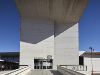 A low-angle photograph of a modern concrete building with a massive cantilevered roof, featuring signage above the entrance that reads "ARTIUM MUSEOA" and "Museo de Arte Contemporáneo del País Vasco".