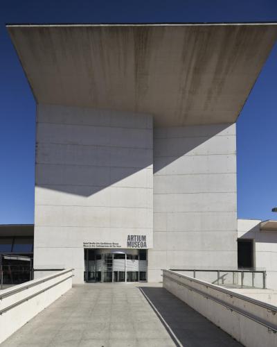 A low-angle photograph of a modern concrete building with a massive cantilevered roof, featuring signage above the entrance that reads "ARTIUM MUSEOA" and "Museo de Arte Contemporáneo del País Vasco".