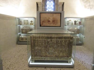 The image shows an interior museum or treasury room containing several ornate, silver reliquary chests and religious artifacts displayed in glass cases, with a large, framed relic mounted on the wall above the central chest.