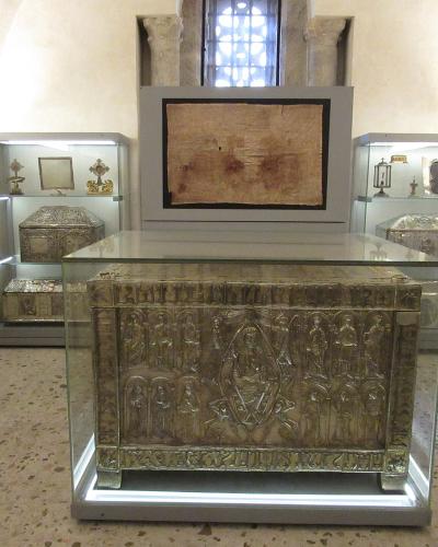 The image shows an interior museum or treasury room containing several ornate, silver reliquary chests and religious artifacts displayed in glass cases, with a large, framed relic mounted on the wall above the central chest.