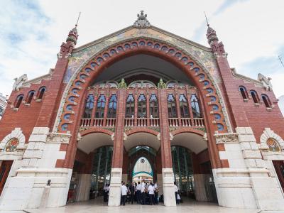 A low-angle photograph of a large, ornate red-brick building with a massive central archway, decorative ceramic tiles, and stone buttresses, with people gathered at the entrance.