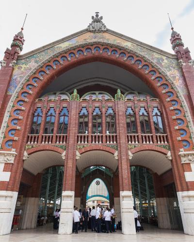 A low-angle photograph of a large, ornate red-brick building with a massive central archway, decorative ceramic tiles, and stone buttresses, with people gathered at the entrance.