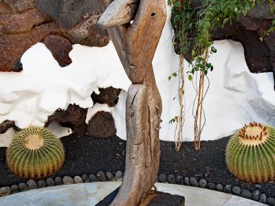 A wooden sculpture of a stylized human figure stands on a volcanic rock base in a courtyard with white-washed walls, black volcanic soil, and barrel cacti.