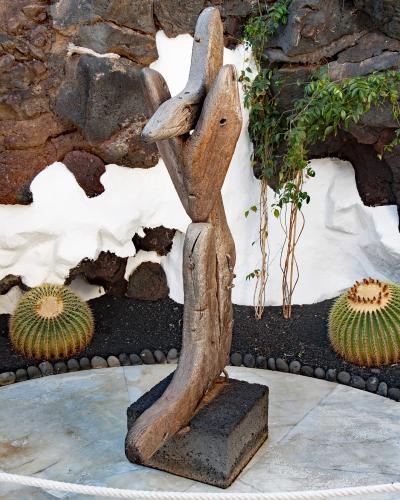 A wooden sculpture of a stylized human figure stands on a volcanic rock base in a courtyard with white-washed walls, black volcanic soil, and barrel cacti.
