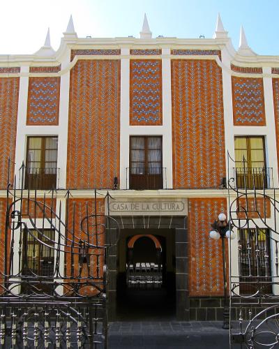 A two-story building with a distinctive orange and white patterned tile facade, white architectural trim, and a central entrance gate with a sign reading "CASA DE LA CULTURA".