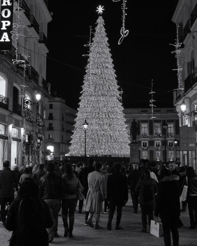 A black and white night photograph of a large, illuminated Christmas tree in a crowded pedestrian plaza, with a vertical "EUROPA" sign on the left building and an equestrian statue in the background.