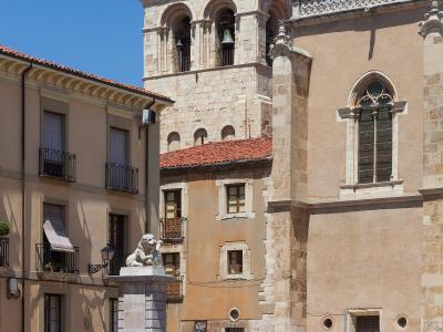 A sunlit plaza featuring a stone lion statue on a pedestal in the foreground, with a historic church tower and adjacent buildings in the background under a clear blue sky.