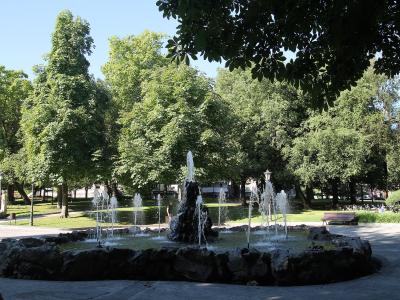 A stone fountain with multiple water jets in a park setting, surrounded by green trees and paved walkways under a clear sky.