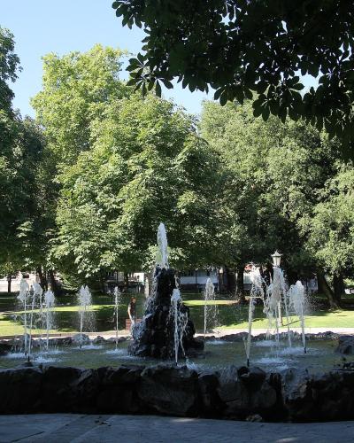 A stone fountain with multiple water jets in a park setting, surrounded by green trees and paved walkways under a clear sky.