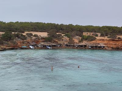 A coastal scene showing a line of rustic wooden boat sheds (varadors) built into a reddish cliffside, with small boats stored underneath, backed by pine trees and a stone building, overlooking turquoise water with swimmers.