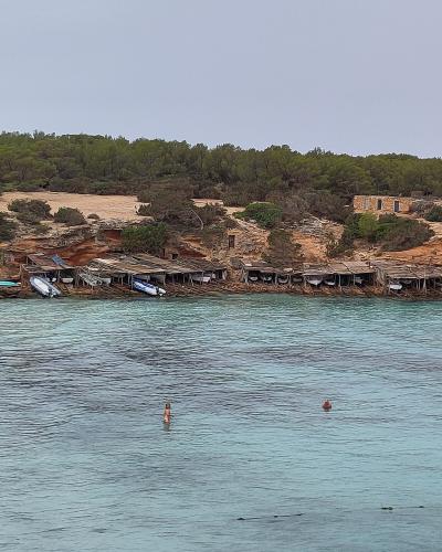A coastal scene showing a line of rustic wooden boat sheds (varadors) built into a reddish cliffside, with small boats stored underneath, backed by pine trees and a stone building, overlooking turquoise water with swimmers.