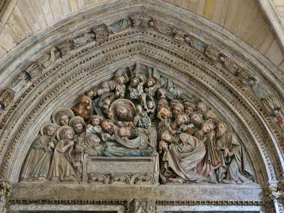 A close-up photograph of a Gothic stone tympanum sculpture depicting the Dormition of the Virgin, featuring the deceased Mary on a bier surrounded by apostles and angels within a decorated arch.