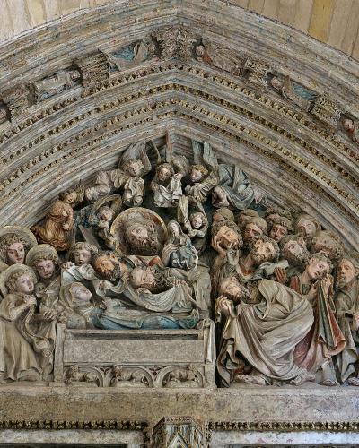 A close-up photograph of a Gothic stone tympanum sculpture depicting the Dormition of the Virgin, featuring the deceased Mary on a bier surrounded by apostles and angels within a decorated arch.