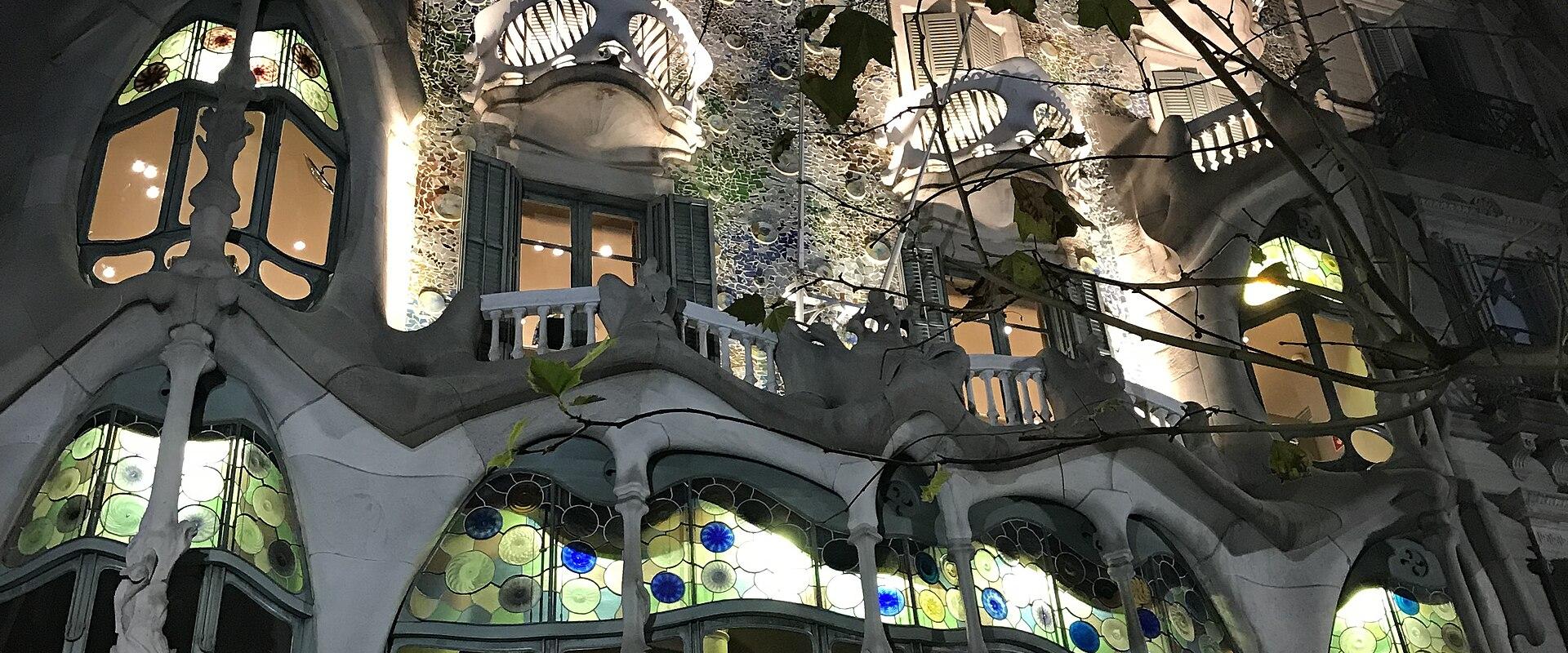 A low-angle photograph of the ornate, bone-like facade of a building with mosaic tiling and skeletal balconies, partially obscured by tree branches, with the text "CASA BATLLÓ" visible in gold lettering above the entrance arches.