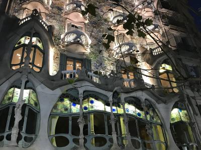A low-angle photograph of the ornate, bone-like facade of a building with mosaic tiling and skeletal balconies, partially obscured by tree branches, with the text "CASA BATLLÓ" visible in gold lettering above the entrance arches.
