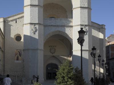 A large, white stone church facade with a distinctive double-arched entrance and two square towers, situated on a paved plaza with planters and lampposts.