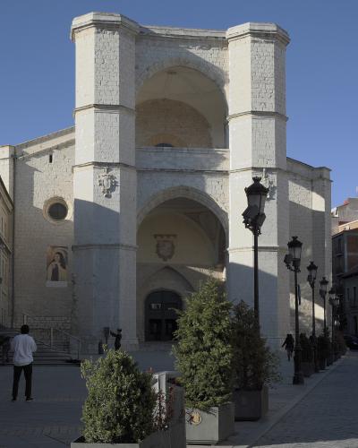 A large, white stone church facade with a distinctive double-arched entrance and two square towers, situated on a paved plaza with planters and lampposts.