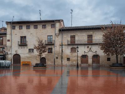 A wet, paved town square featuring a large, two-story stone building with a prominent corner tower, arched ground-floor doorways, and a row of iron chains hanging across the facade.