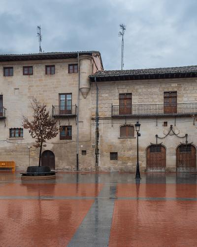 A wet, paved town square featuring a large, two-story stone building with a prominent corner tower, arched ground-floor doorways, and a row of iron chains hanging across the facade.