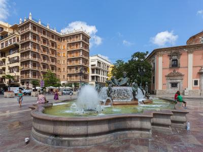 A large public square featuring a prominent fountain with bronze statues of figures and horses, flanked by a multi-story residential building on the left and a pinkish church facade on the right under a blue sky.