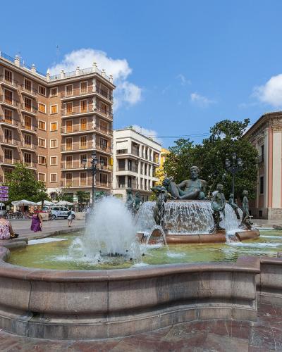 A large public square featuring a prominent fountain with bronze statues of figures and horses, flanked by a multi-story residential building on the left and a pinkish church facade on the right under a blue sky.