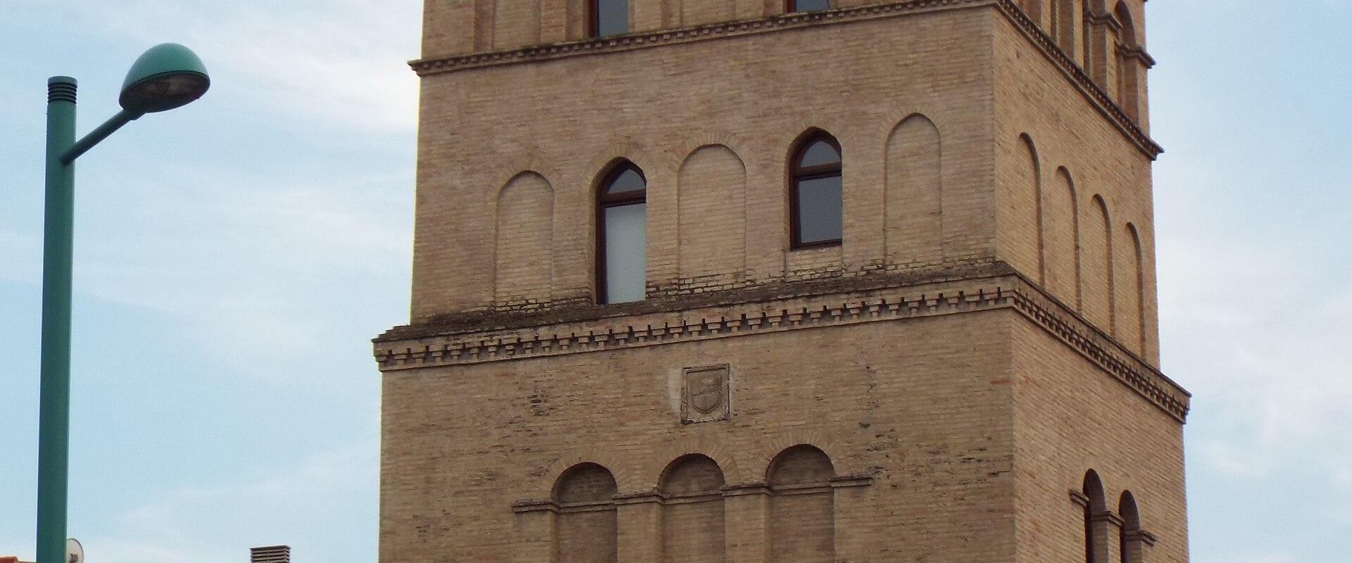 A tall, four-story tower constructed of light-colored brick, featuring arched windows, a decorative cornice, and a small finial on the roof, with a red banner attached to its lower right side.