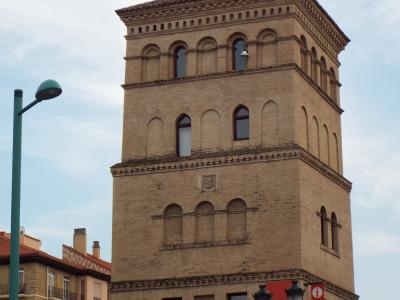 A tall, four-story tower constructed of light-colored brick, featuring arched windows, a decorative cornice, and a small finial on the roof, with a red banner attached to its lower right side.
