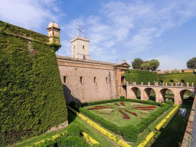 A stone fortress with high walls covered in ivy, a central tower flying flags, and formal gardens with geometric flowerbeds in the foreground.