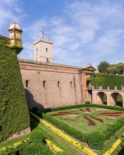 A stone fortress with high walls covered in ivy, a central tower flying flags, and formal gardens with geometric flowerbeds in the foreground.