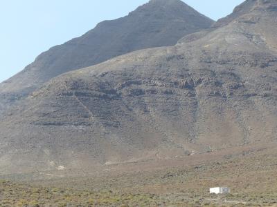 A view of arid, rocky volcanic mountains with sparse scrub vegetation in the foreground and a small white building at the base.