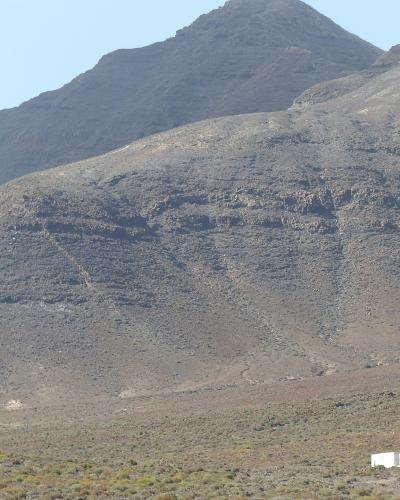 A view of arid, rocky volcanic mountains with sparse scrub vegetation in the foreground and a small white building at the base.