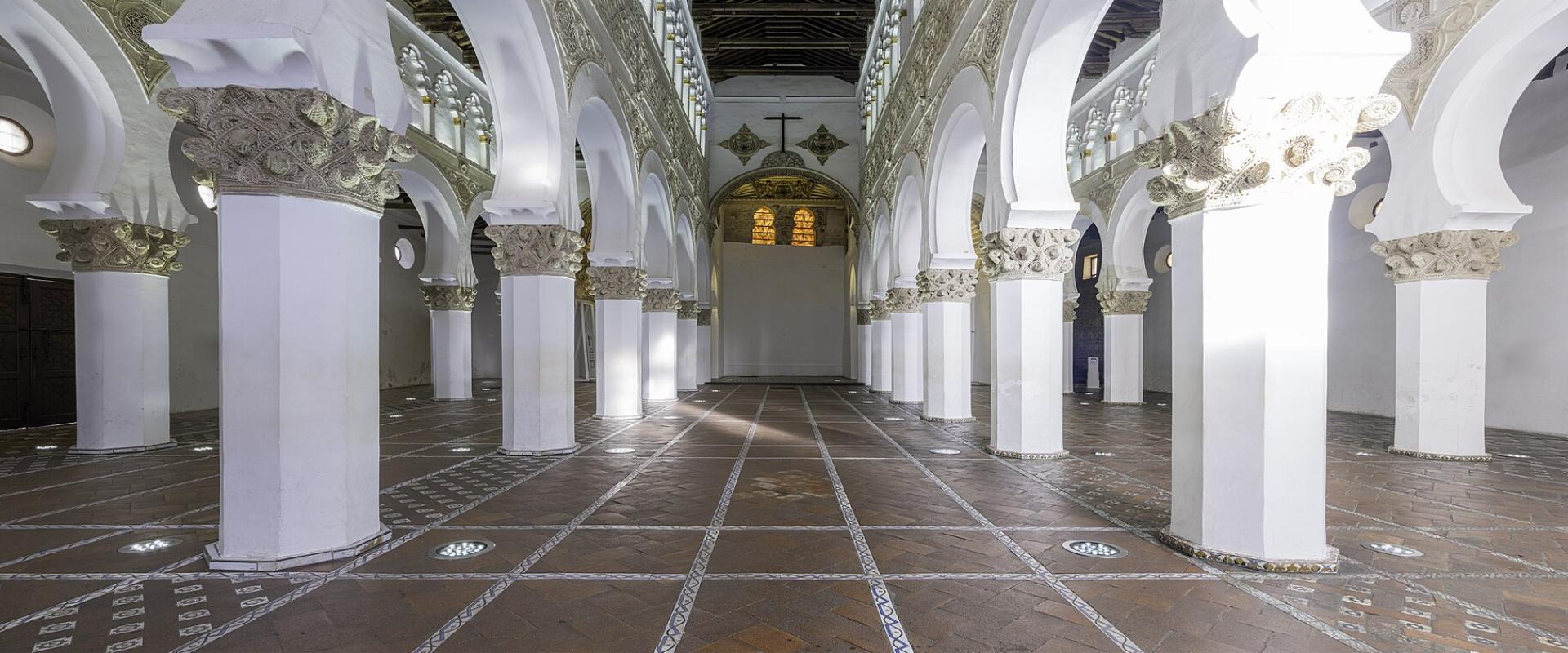 A panoramic interior view of a large hall featuring rows of white octagonal columns supporting double-tiered horseshoe arches with ornate capitals, leading to an apse with a Christian cross and iconography.