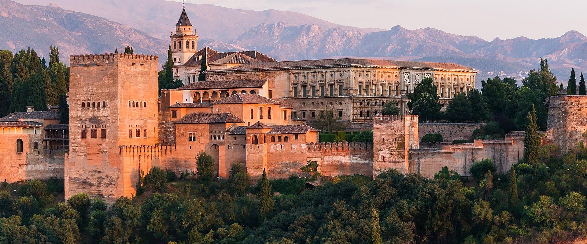 A panoramic view of the Alhambra fortress complex in Granada, featuring the Palace of Charles V with its distinctive circular courtyard and Renaissance facade, alongside the Alcazaba towers and the Cathedral spire, set against the Sierra Nevada mount