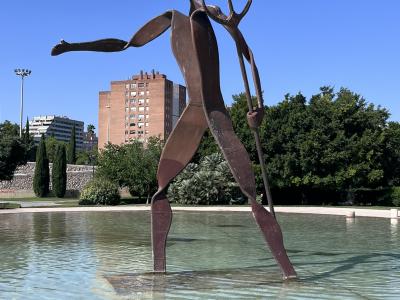 A large, abstract, rust-colored metal sculpture of a humanoid figure holding a trident, standing in a shallow pool of water with trees and a brick apartment building in the background.