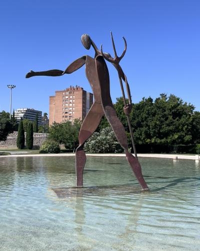A large, abstract, rust-colored metal sculpture of a humanoid figure holding a trident, standing in a shallow pool of water with trees and a brick apartment building in the background.