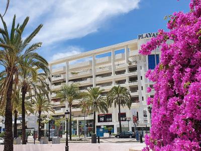 A street-level view of a white multi-story building with a large pink bougainvillea bush in the foreground and palm trees lining the sidewalk.