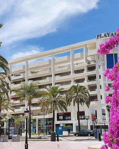 A street-level view of a white multi-story building with a large pink bougainvillea bush in the foreground and palm trees lining the sidewalk.