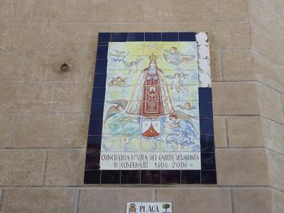 A close-up of a tiled religious mural and a street sign mounted on a beige stone wall, depicting the Virgin Mary with angels and text reading "CONFRARIA NTRA SRA DEL CARME DE VALÈNCIA" and "PLAÇA DEL CARME".
