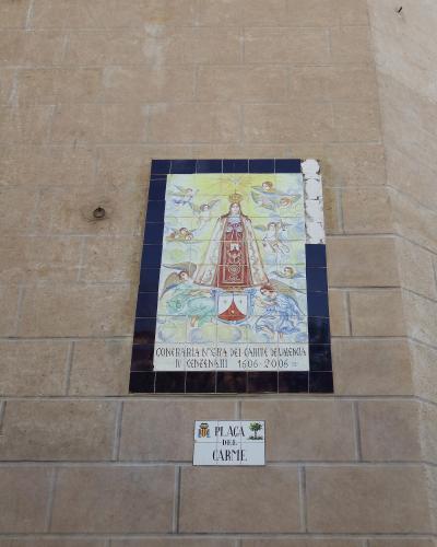 A close-up of a tiled religious mural and a street sign mounted on a beige stone wall, depicting the Virgin Mary with angels and text reading "CONFRARIA NTRA SRA DEL CARME DE VALÈNCIA" and "PLAÇA DEL CARME".