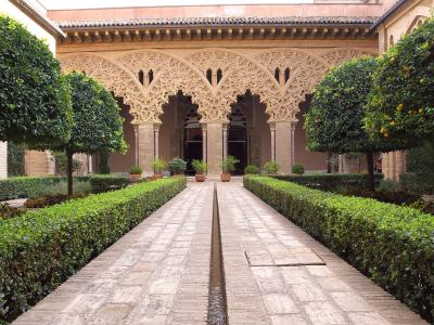 A courtyard garden with a central stone water channel, flanked by manicured hedges and orange trees, leading to an ornate Moorish-style arcade with intricate stucco work and horseshoe arches.