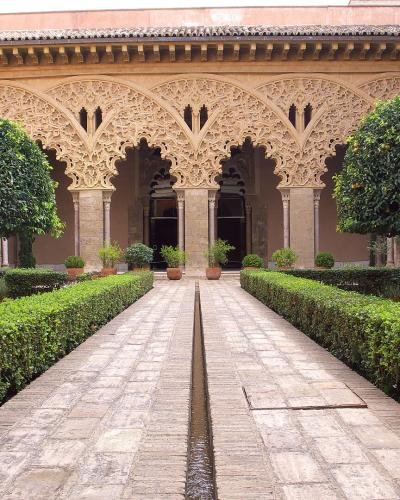A courtyard garden with a central stone water channel, flanked by manicured hedges and orange trees, leading to an ornate Moorish-style arcade with intricate stucco work and horseshoe arches.