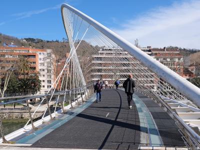 A white, cable-stayed pedestrian bridge with a curved, white arch structure, set against a blue sky, with people walking on the path and urban buildings visible in the background.