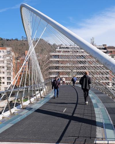 A white, cable-stayed pedestrian bridge with a curved, white arch structure, set against a blue sky, with people walking on the path and urban buildings visible in the background.