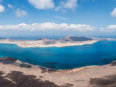 A panoramic view from a high, arid volcanic vantage point looking across turquoise water towards the island of La Graciosa, with its distinctive coastline and settlement visible in the distance.