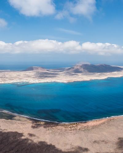 A panoramic view from a high, arid volcanic vantage point looking across turquoise water towards the island of La Graciosa, with its distinctive coastline and settlement visible in the distance.