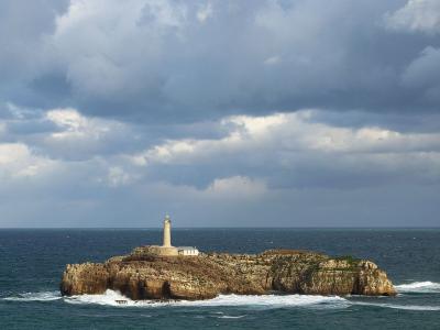 A conical white lighthouse situated on a small, rocky island surrounded by the sea under a cloudy sky.