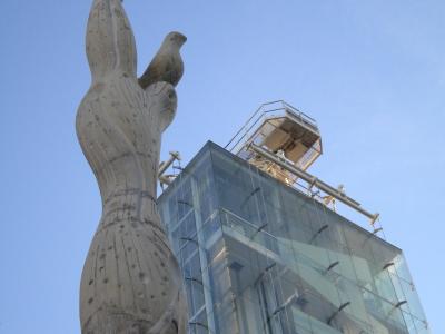 A low-angle shot of a tall, abstract stone sculpture featuring a bird motif, set against a modern glass building with a metal observation platform on its roof under a clear blue sky.
