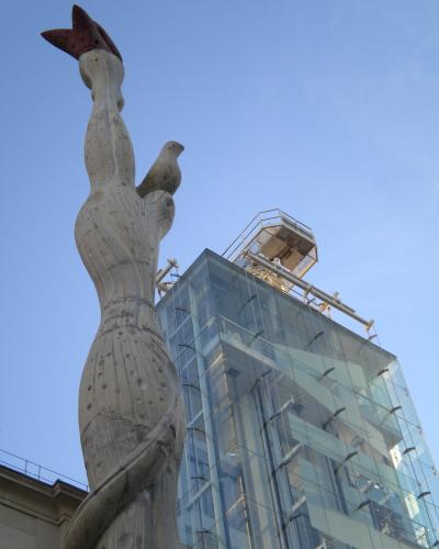 A low-angle shot of a tall, abstract stone sculpture featuring a bird motif, set against a modern glass building with a metal observation platform on its roof under a clear blue sky.