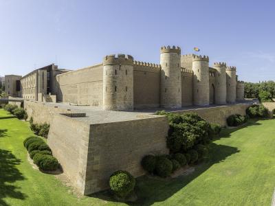 A large medieval fortress with crenellated stone walls, round towers, and a flag flying from the highest tower, surrounded by manicured green lawns and hedges under a clear blue sky.